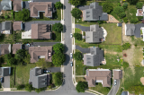 Aerial top-down view of a suburban neighborhood with single-family houses, green lawns, driveways, and trees lining a grid of residential streets, with a few cars parked along the road.