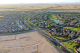 Aerial view of a large suburban neighborhood in a desert landscape, with curving residential streets, single-family homes, a small lake with a fountain, and a golf course, bordered by open undeveloped land and distant mountains.