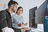 Photo of two young professionals looking at lines of code on two monitors.